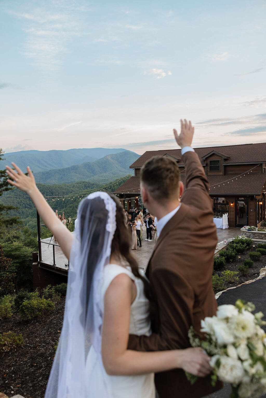 Bride and groom waving to their guests from above with the wedding venue and Smoky Mountains visible below.