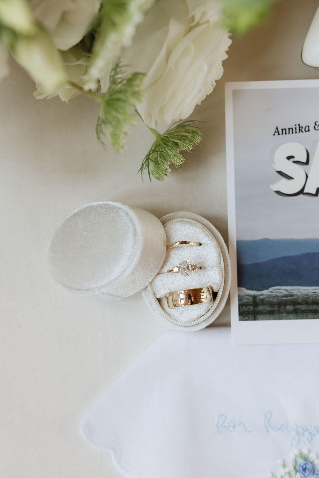 Wedding rings displayed in a white velvet ring box beside white flowers and wedding stationery.