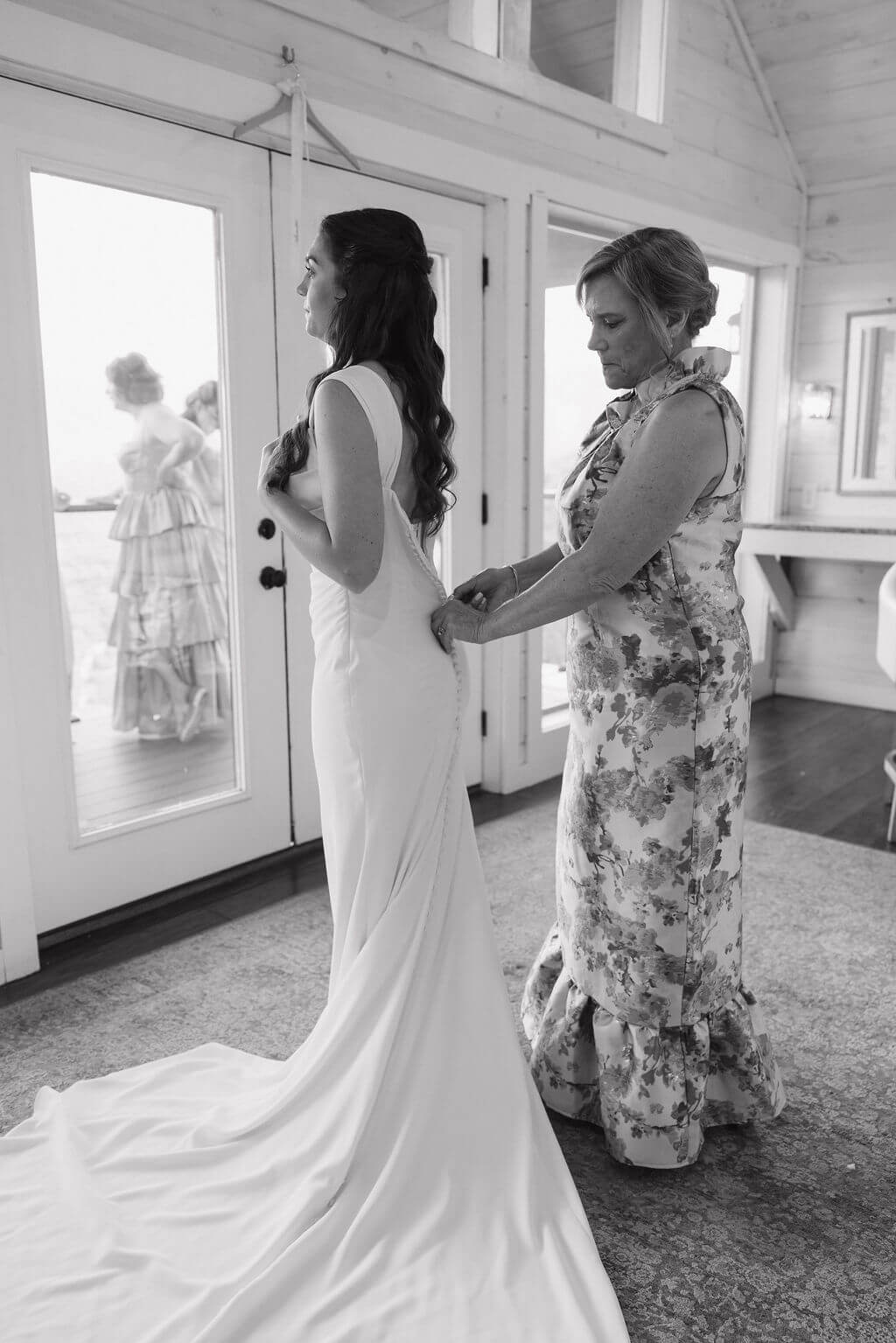 Black-and-white photo of a woman helping the bride button the back of her wedding dress while getting ready.