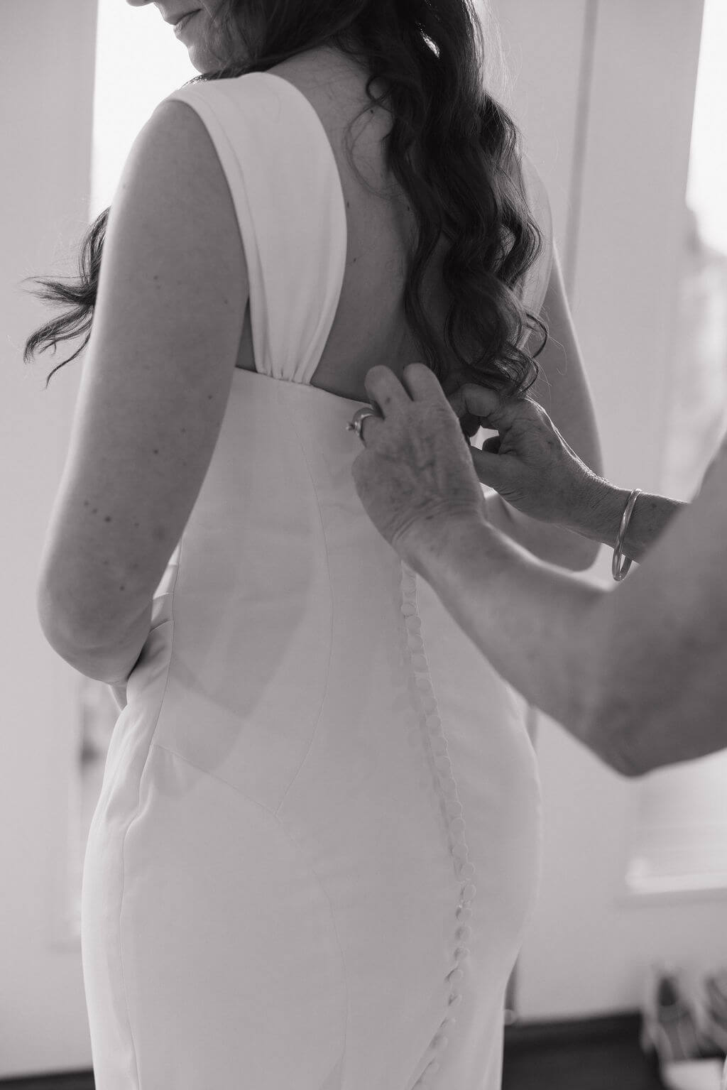 Black-and-white close-up of someone fastening the buttons on the back of the bride’s wedding dress.
