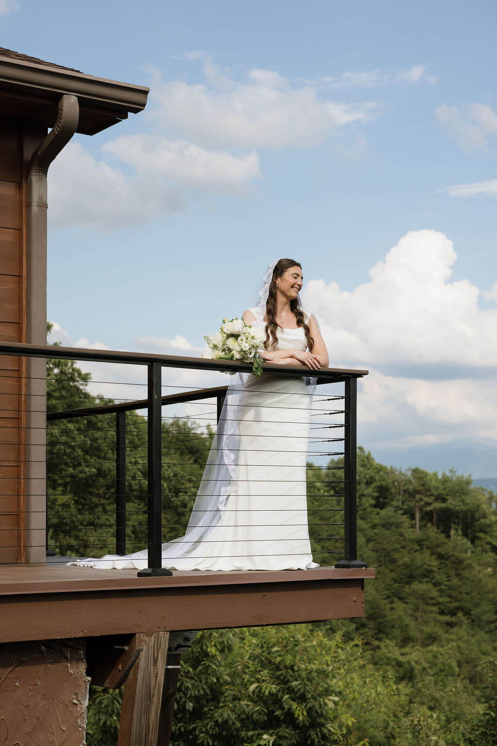 Bride standing on a wooden balcony holding a white bouquet with blue sky and mountain views behind her.