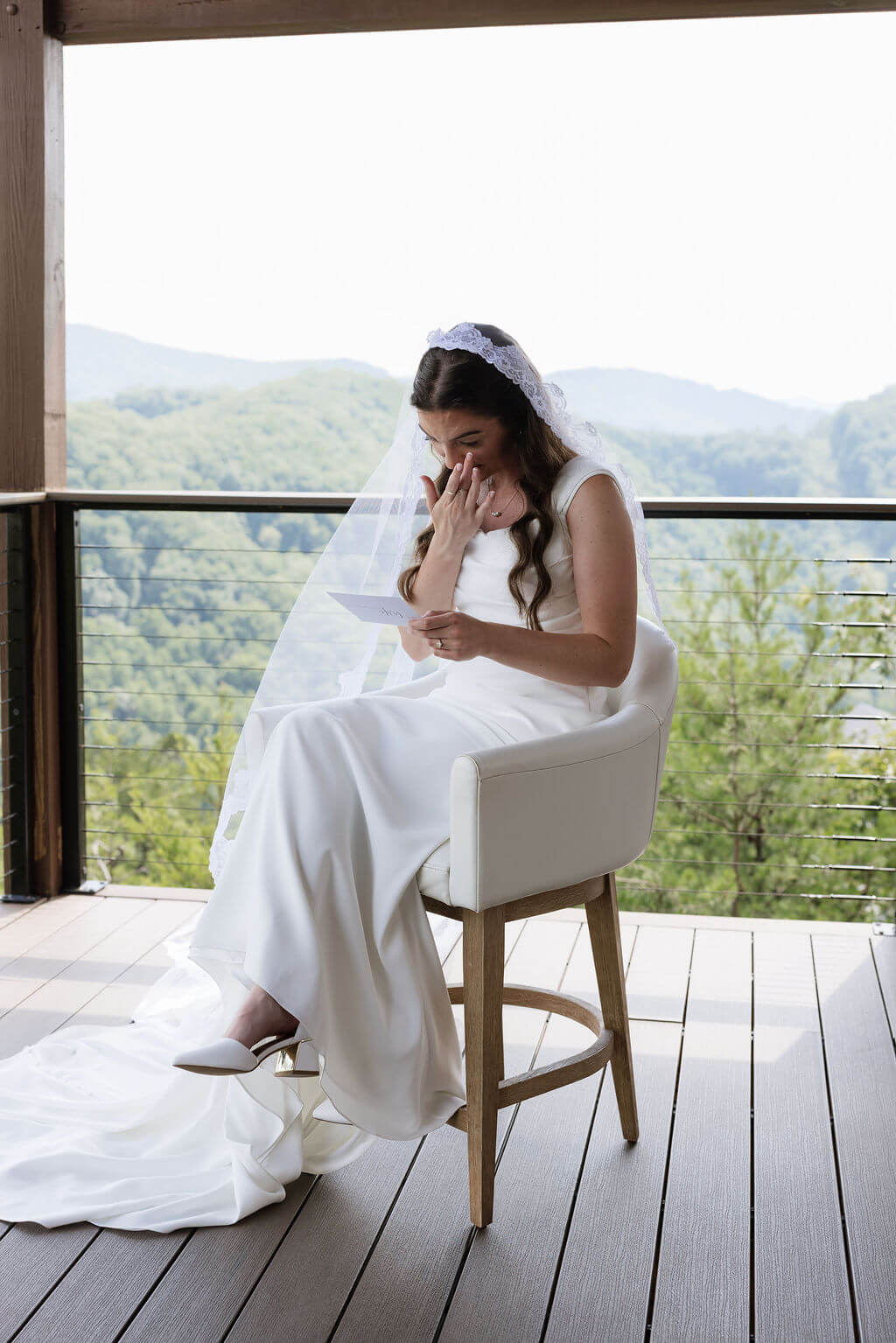 Bride sitting on a balcony reading a letter with sweeping green mountain views behind her.