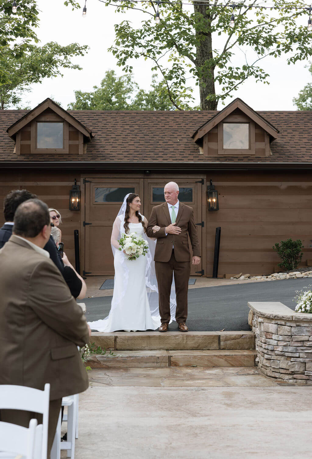 Bride walking toward the outdoor ceremony with an older man as seated guests look on.