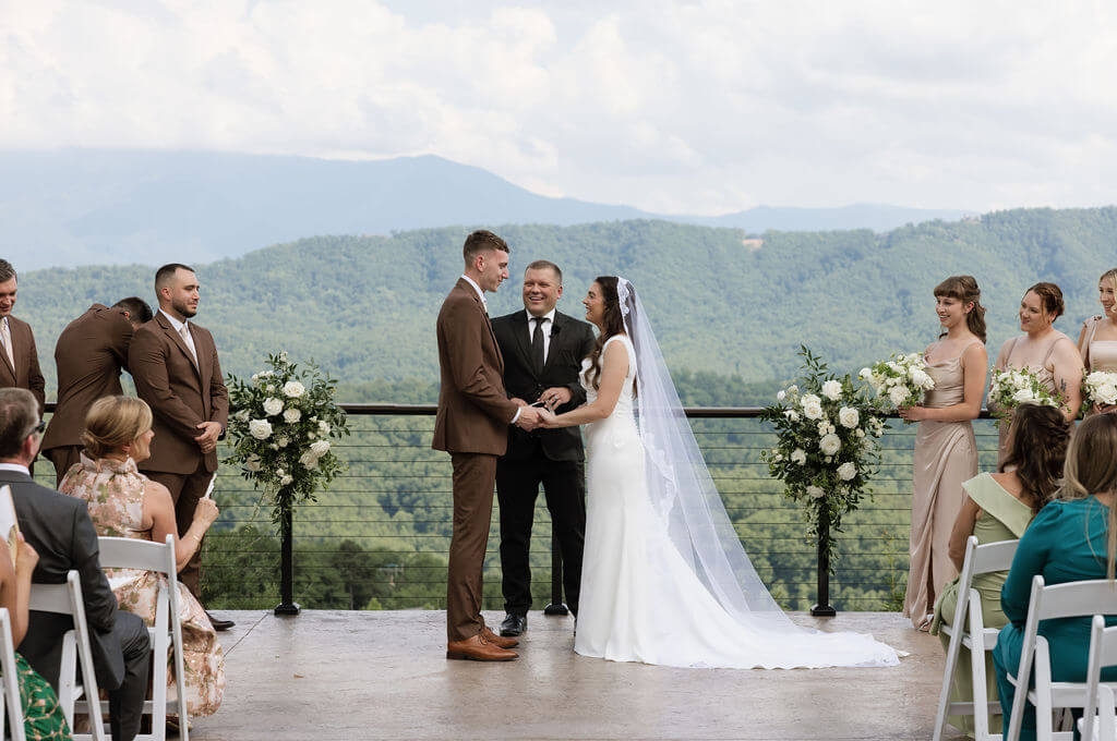 Bride and groom holding hands during their outdoor wedding ceremony with the officiant, wedding party, and mountains behind them.