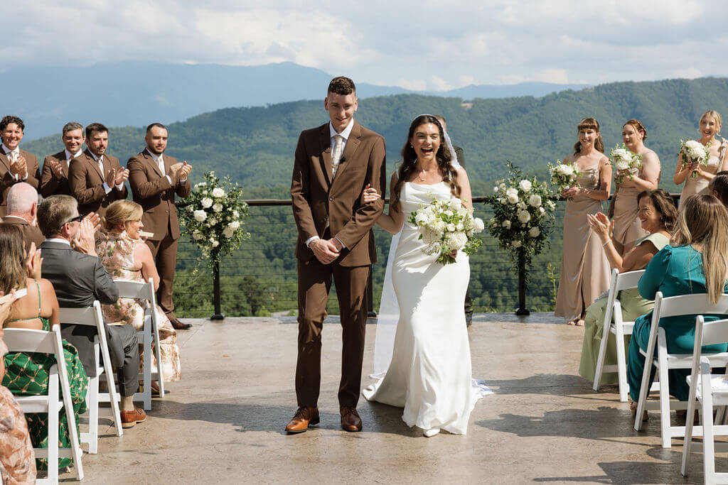 Newlyweds smiling as they walk back down the aisle while their wedding party and guests applaud against a mountain backdrop.