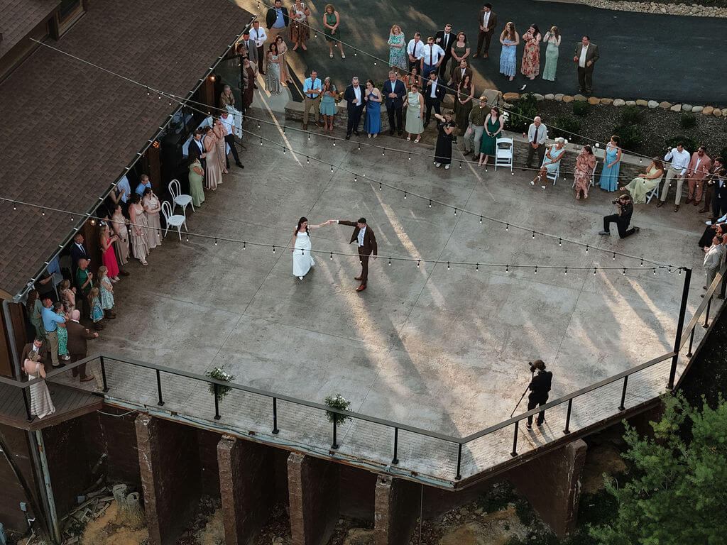 Aerial view of the bride and groom dancing together on an outdoor terrace while wedding guests gather around them under string lights.