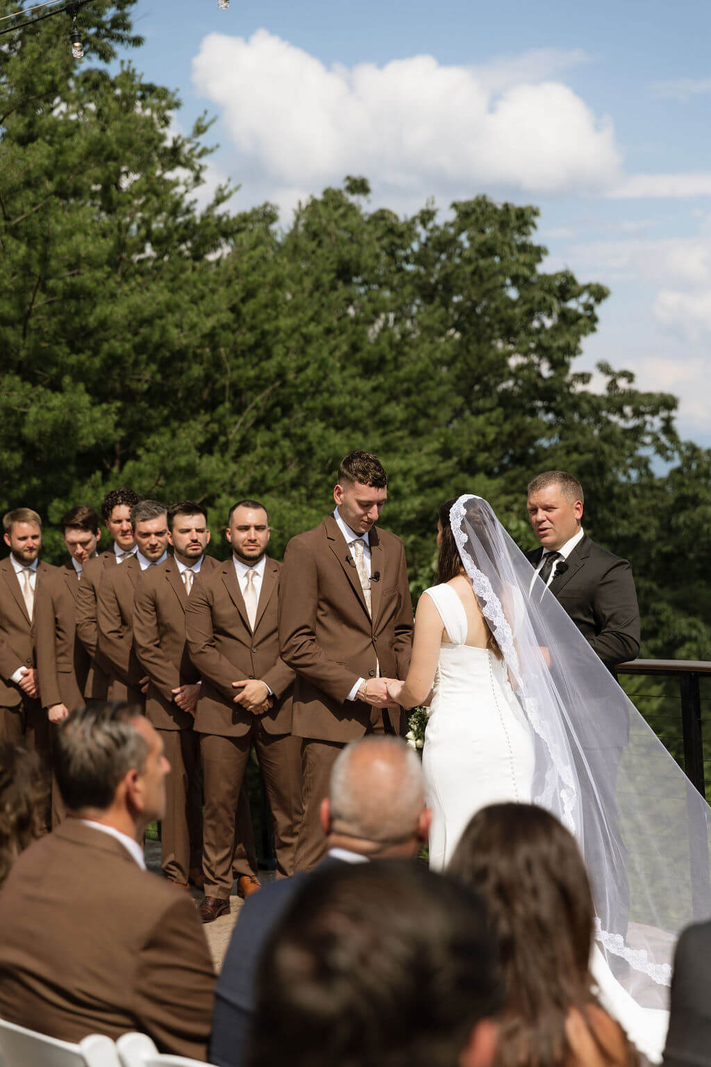 Bride and groom exchanging rings during their outdoor wedding ceremony while their wedding party stands behind them.