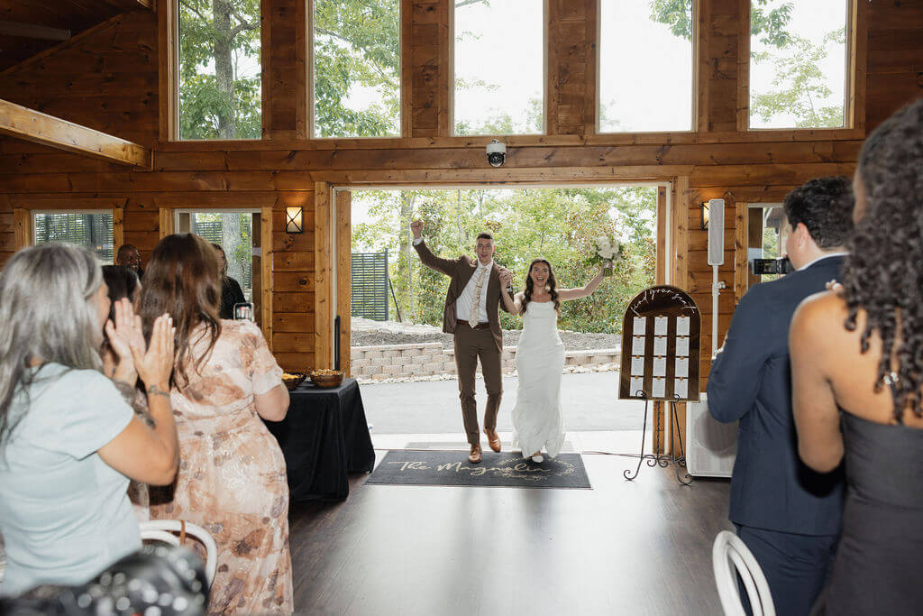 Newlyweds entering their reception with their arms raised while guests applaud inside the wooden mountain venue.