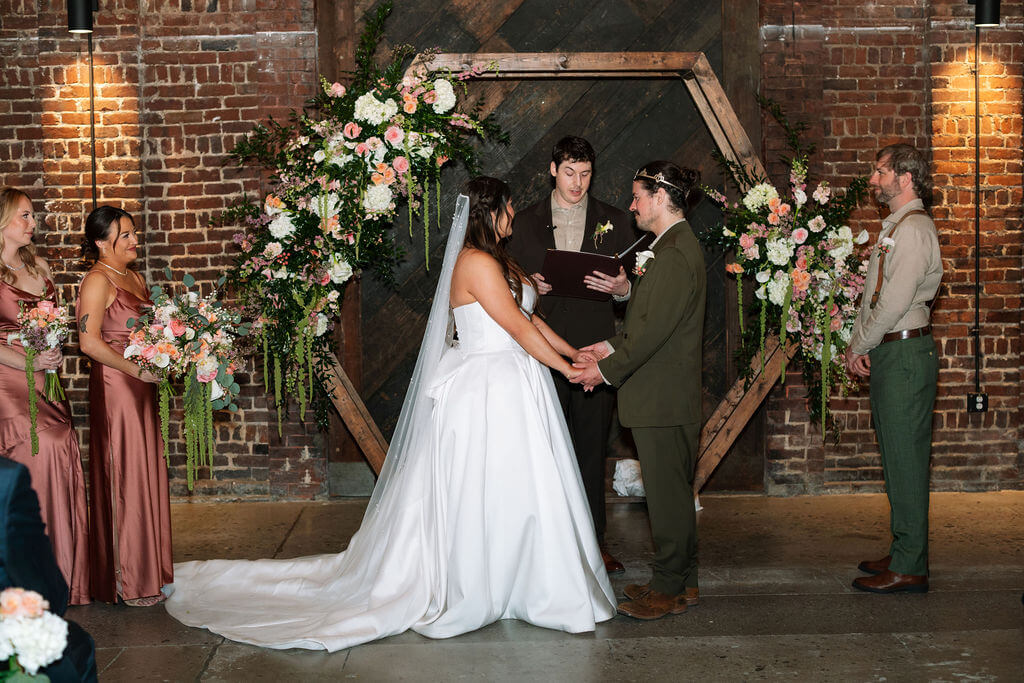 Bride in a strapless satin gown and cathedral veil holding hands with the groom in an olive suit during their ceremony in front of a floral hexagon arch and exposed brick walls at Jackson Terminal
