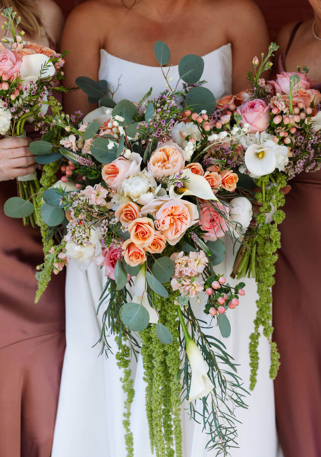 Close-up of a cascading bridal bouquet with peach garden roses, pink roses, white calla lilies, eucalyptus, and trailing green amaranthus against a bridesmaid's mauve satin dress