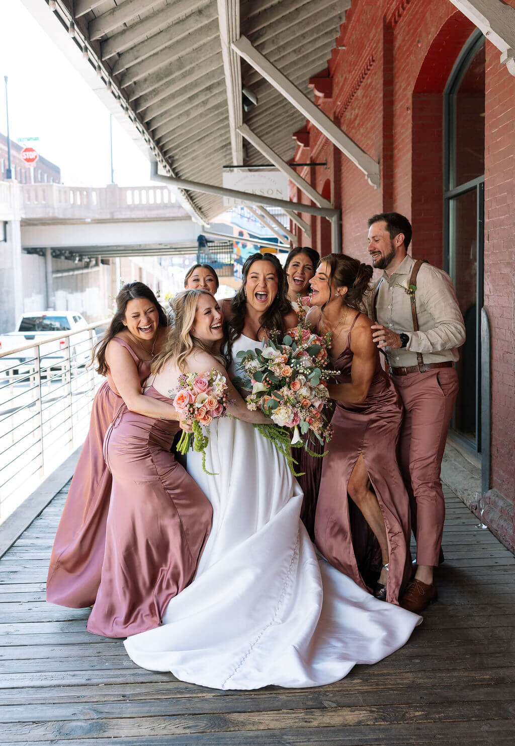 Bride laughing with her bridesmaids in mauve satin dresses and a groomsman in suspenders on the covered wooden platform at Jackson Terminal in Knoxville
