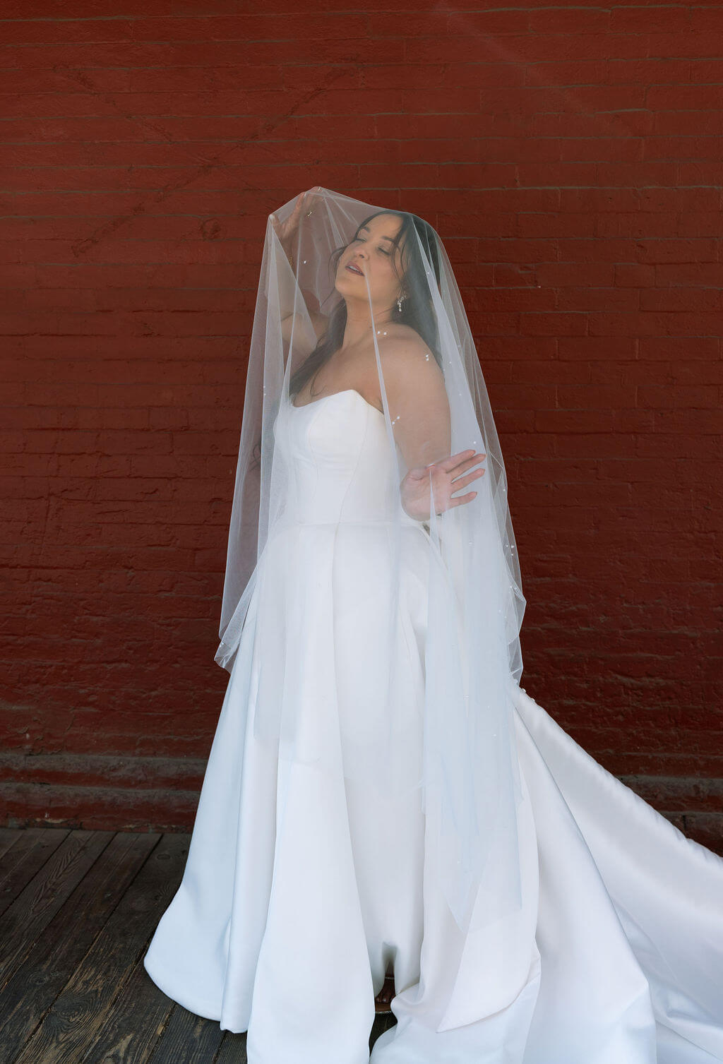 Bride standing in front of a deep red wall with her sheer veil draped over her head and shoulders