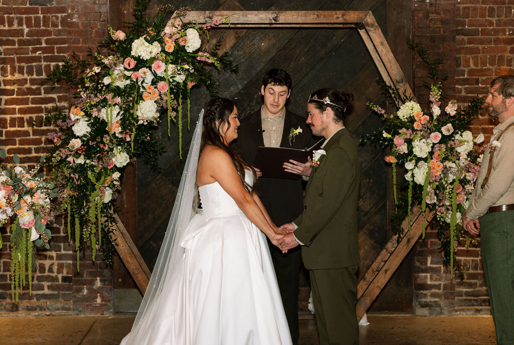 Wide view of the ceremony with the bride and groom holding hands in front of a floral hexagon arch, the officiant reading from a black folder, and a groomsman standing nearby