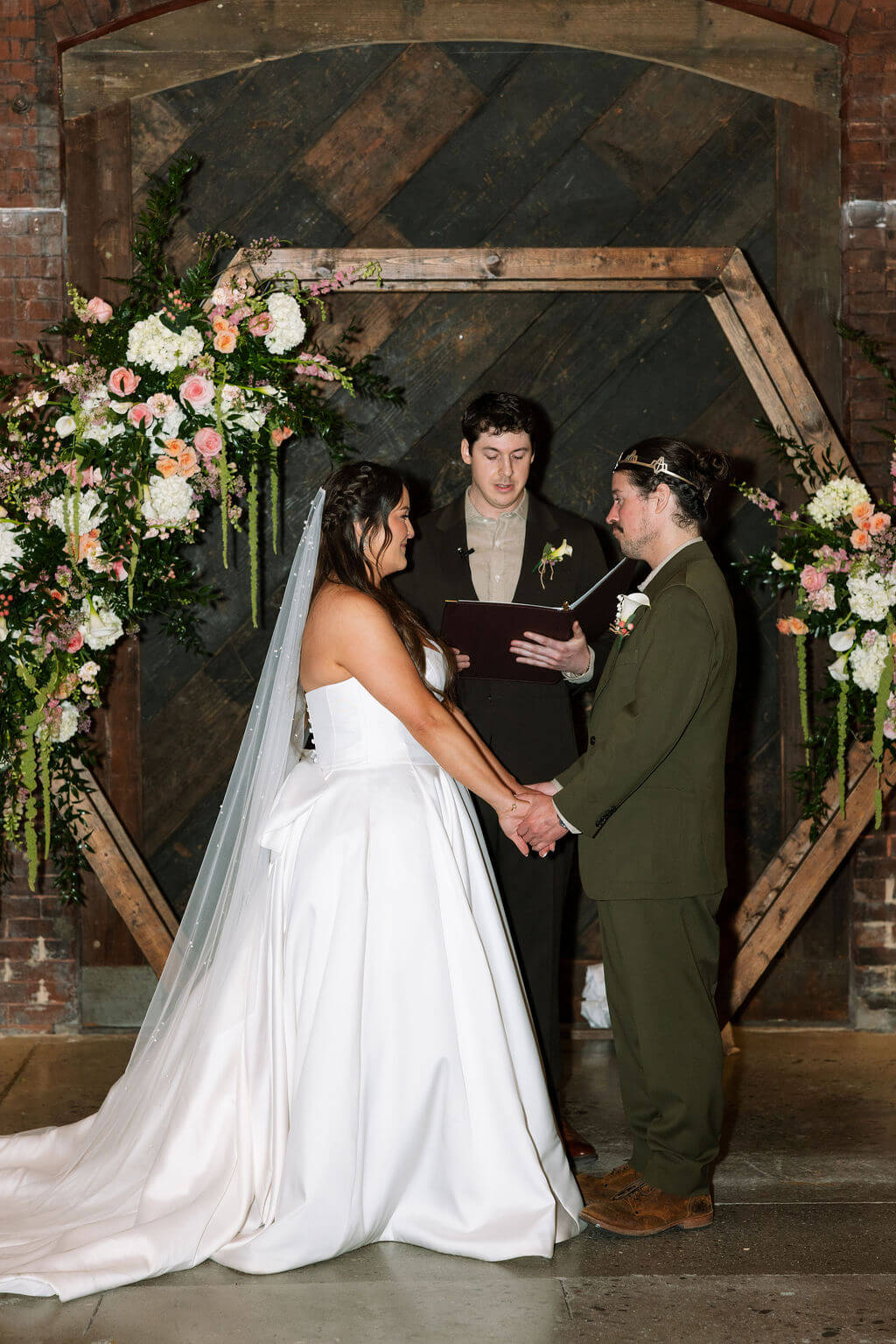 Bride and groom holding hands at the altar under the wooden hexagon arch as the officiant leads the ceremony