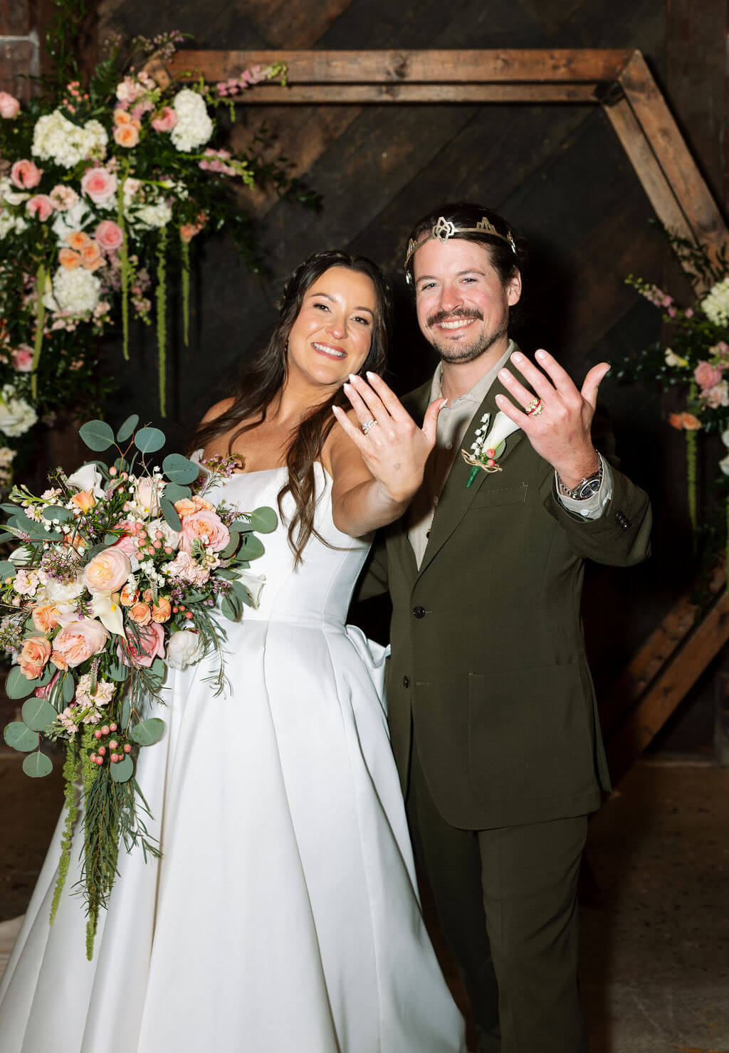 Bride and groom smiling and showing off their wedding rings in front of a wooden hexagon arch decorated with peach and pink florals