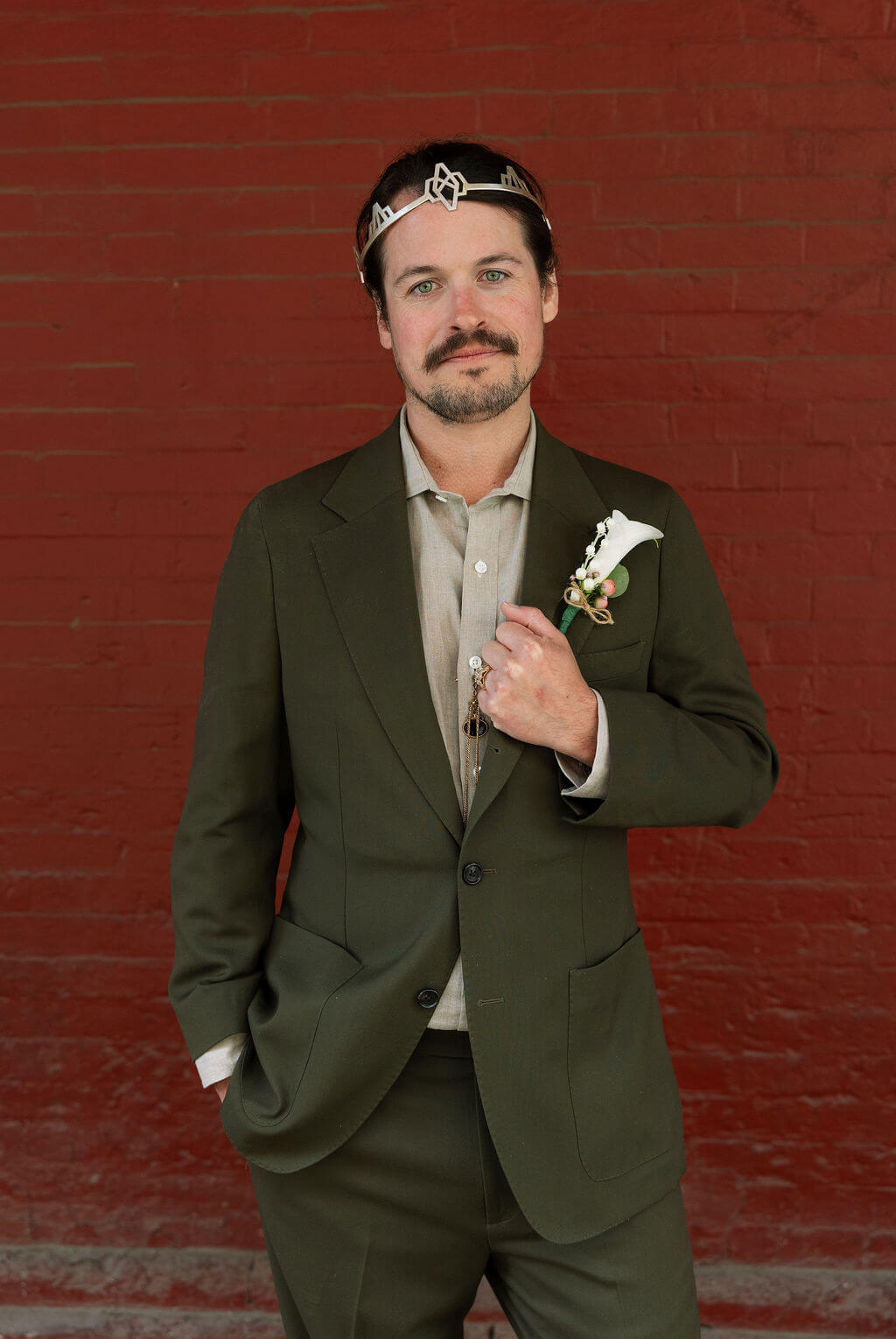 Groom in an olive suit and silver crown posing with his hand on his lapel in front of a dark red wall