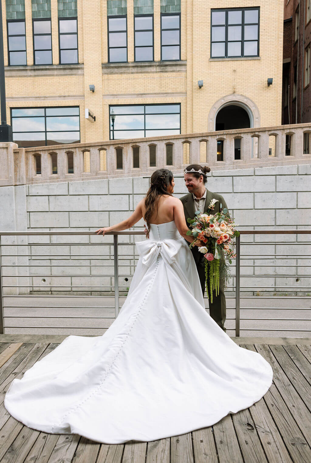 Bride and groom leaning against a metal railing on a wooden deck, showing the oversized bow and long train on the back of the bride's satin wedding gown