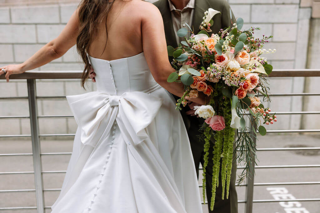 Back view of the bride's strapless satin gown with an oversized bow and button detail as the groom holds her cascading bouquet by a metal railing
