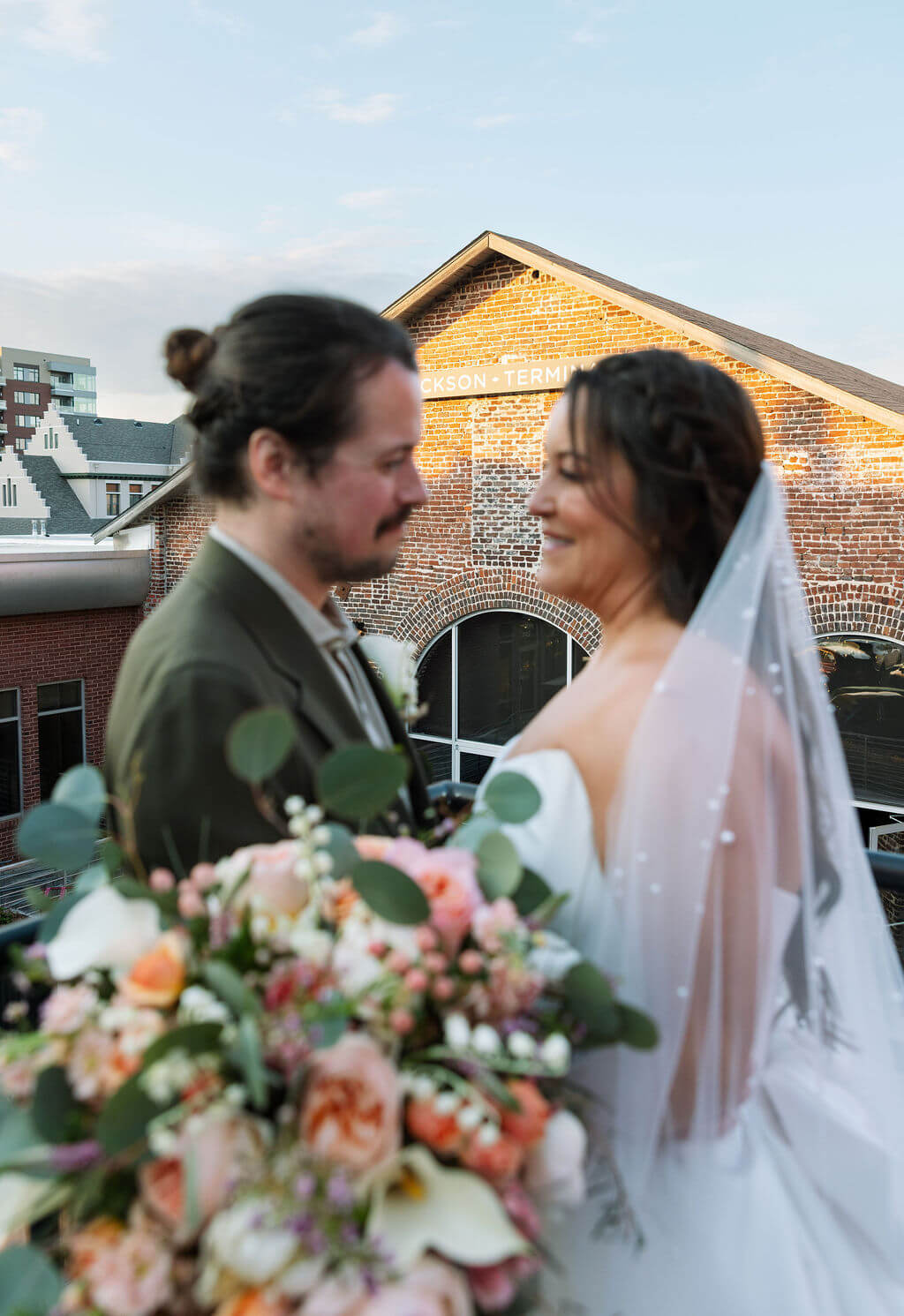 Bride and groom gazing at each other with the brick Jackson Terminal building behind them and a peach and pink bouquet in the foreground