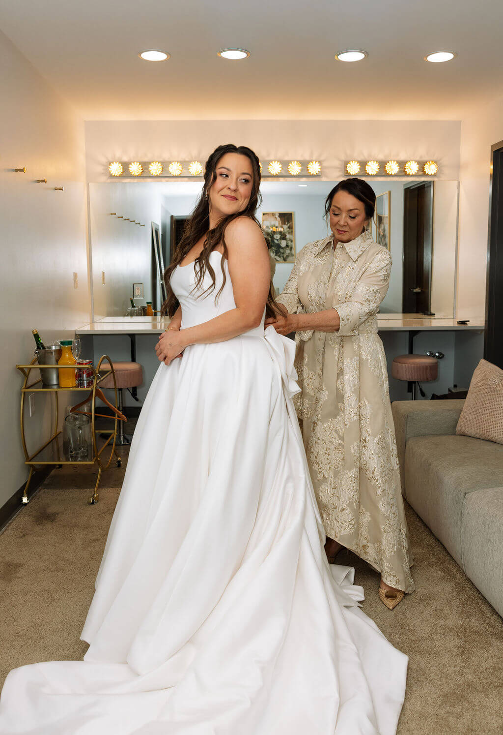 Bride stands in her wedding dress while a family member helps adjust her gown inside a bright bridal suite.