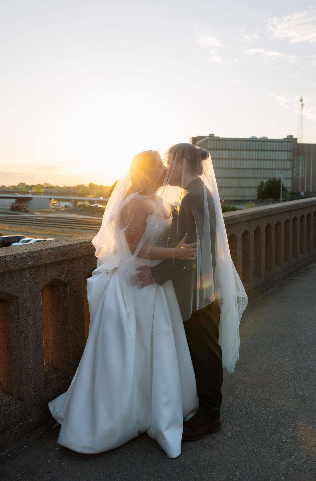 Bride and groom kissing at sunset on a bridge overlooking downtown Knoxville with the bride's veil draped around them