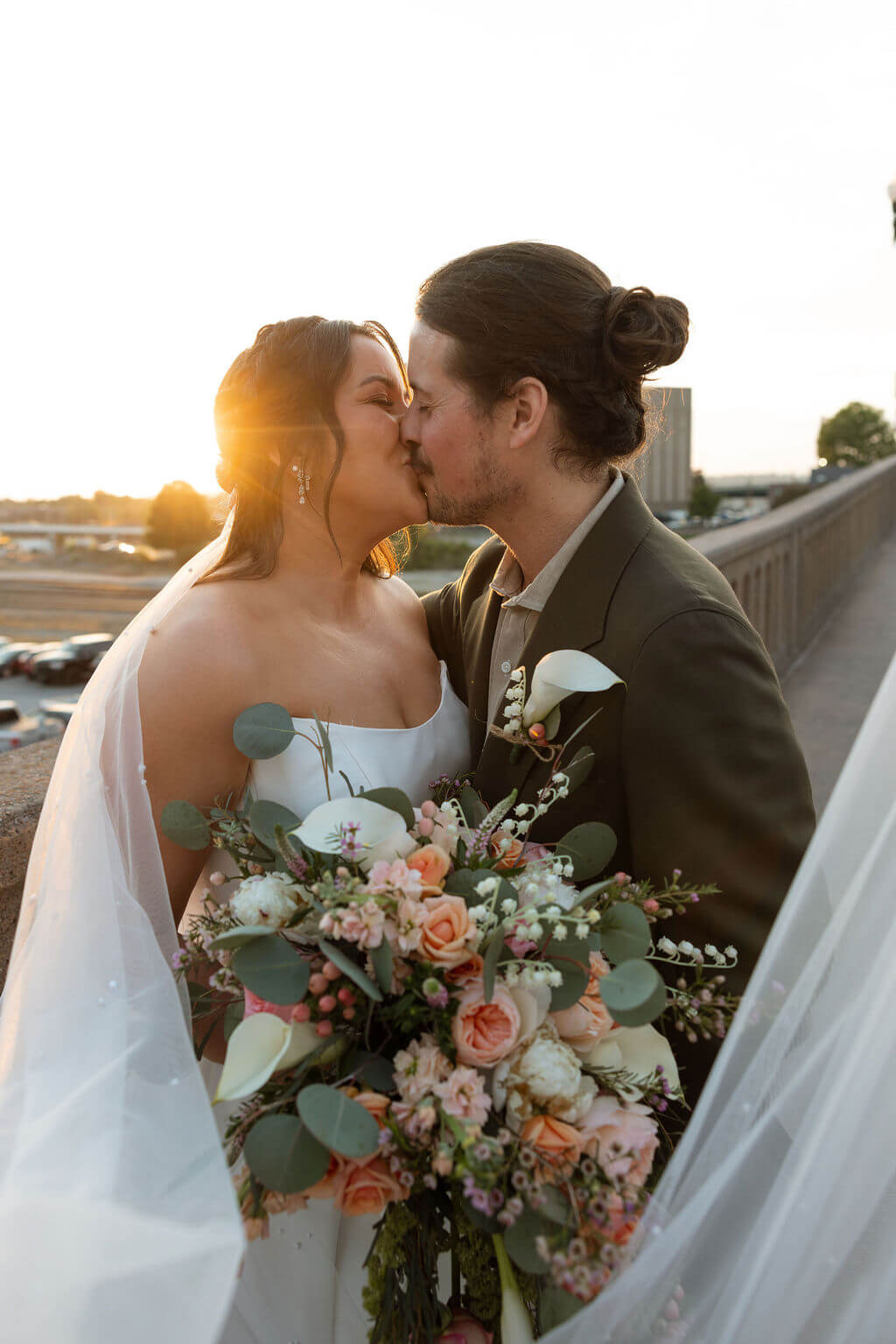 Bride and groom kissing at golden hour as the sun flares behind them, the bride's bouquet with calla lilies in front