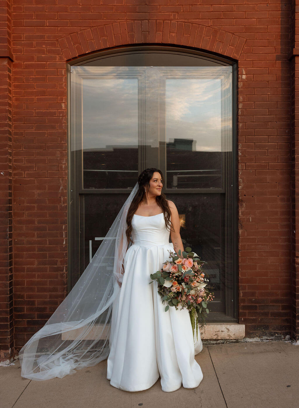 Bride standing with her veil and bouquet in front of an arched window set into a red brick wall