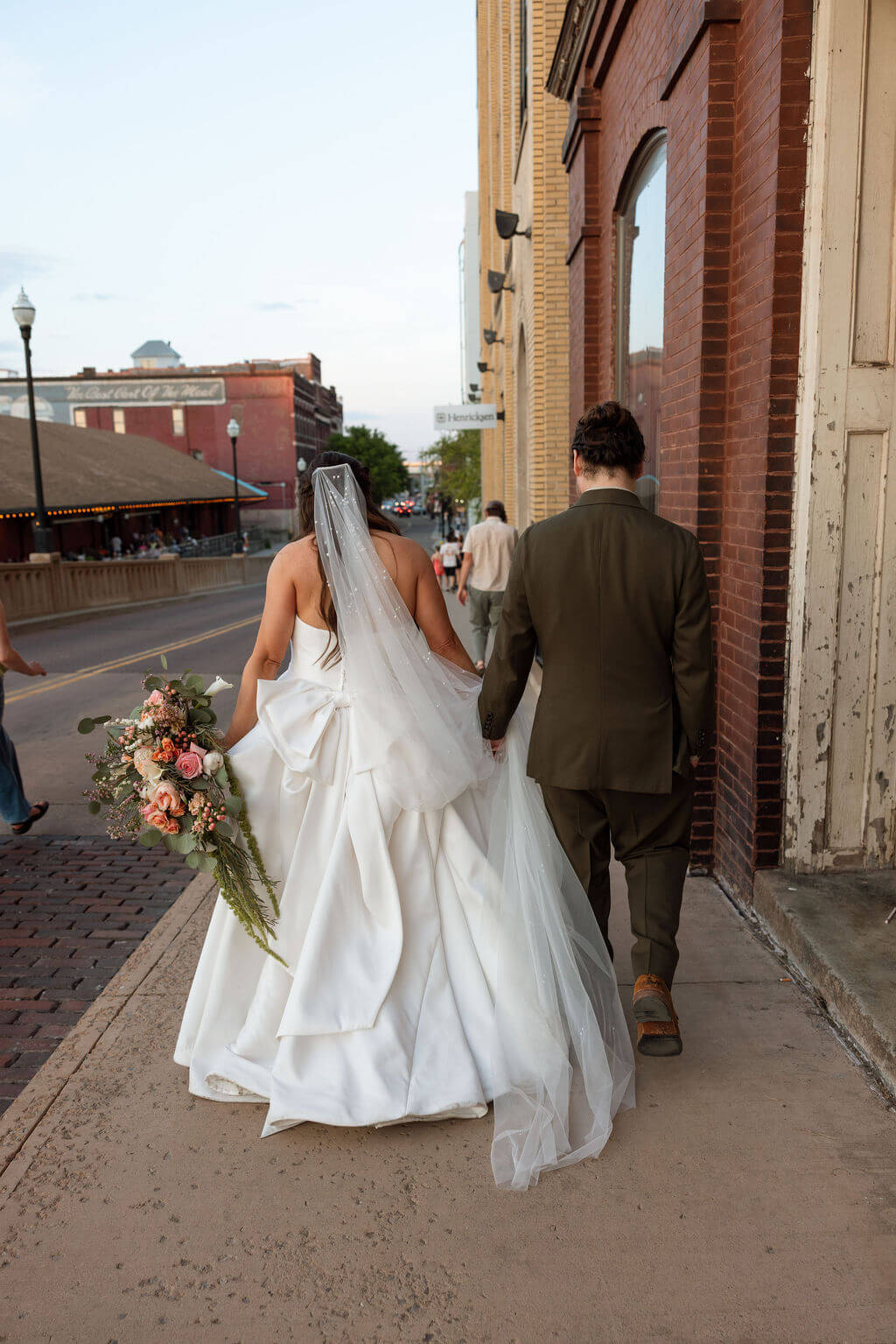Bride and groom walking away down a sidewalk lined with historic brick buildings, the bride's veil trailing behind her
