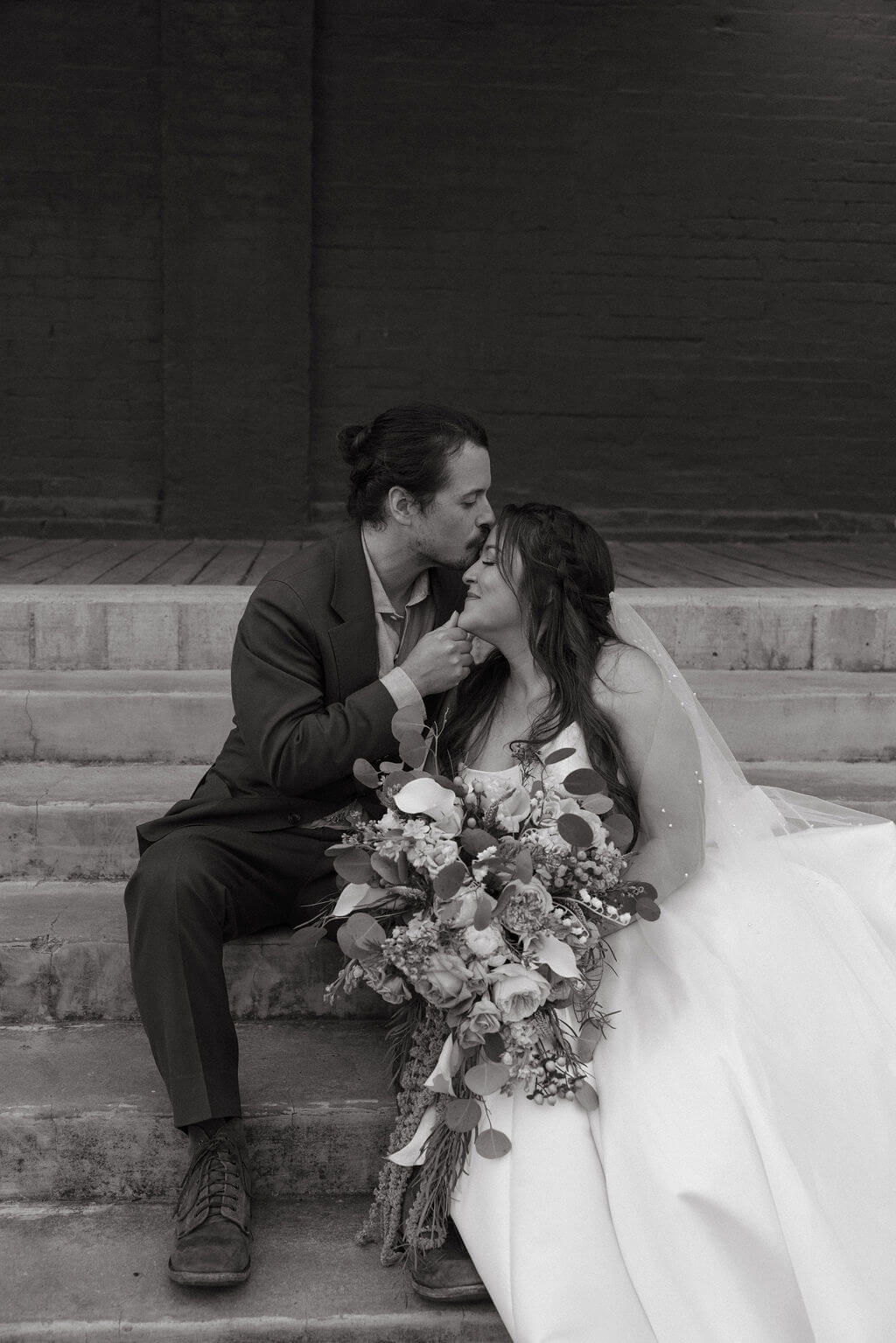 Black and white photo of the groom kissing the bride's forehead as they sit together on concrete steps