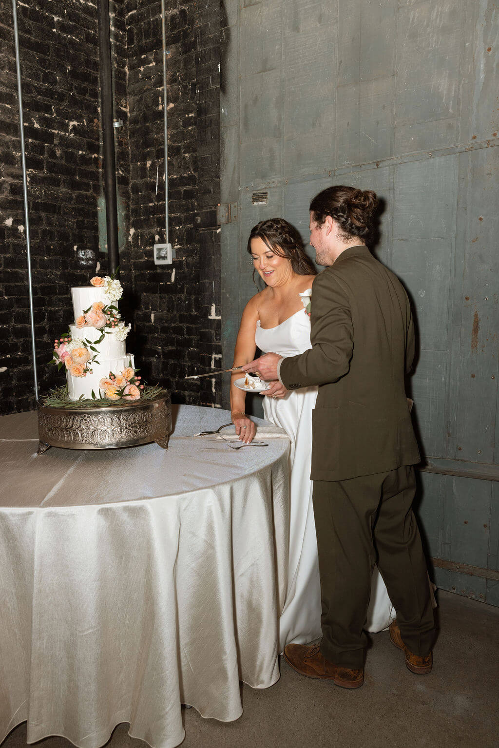 Bride and groom cutting a white tiered wedding cake decorated with fresh flowers beside a brick wall