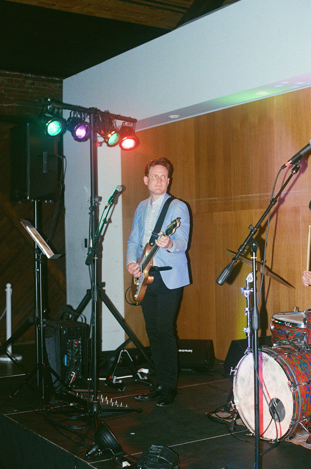 Band guitarist performing on stage under colorful lights with a drum kit beside him during the wedding reception
