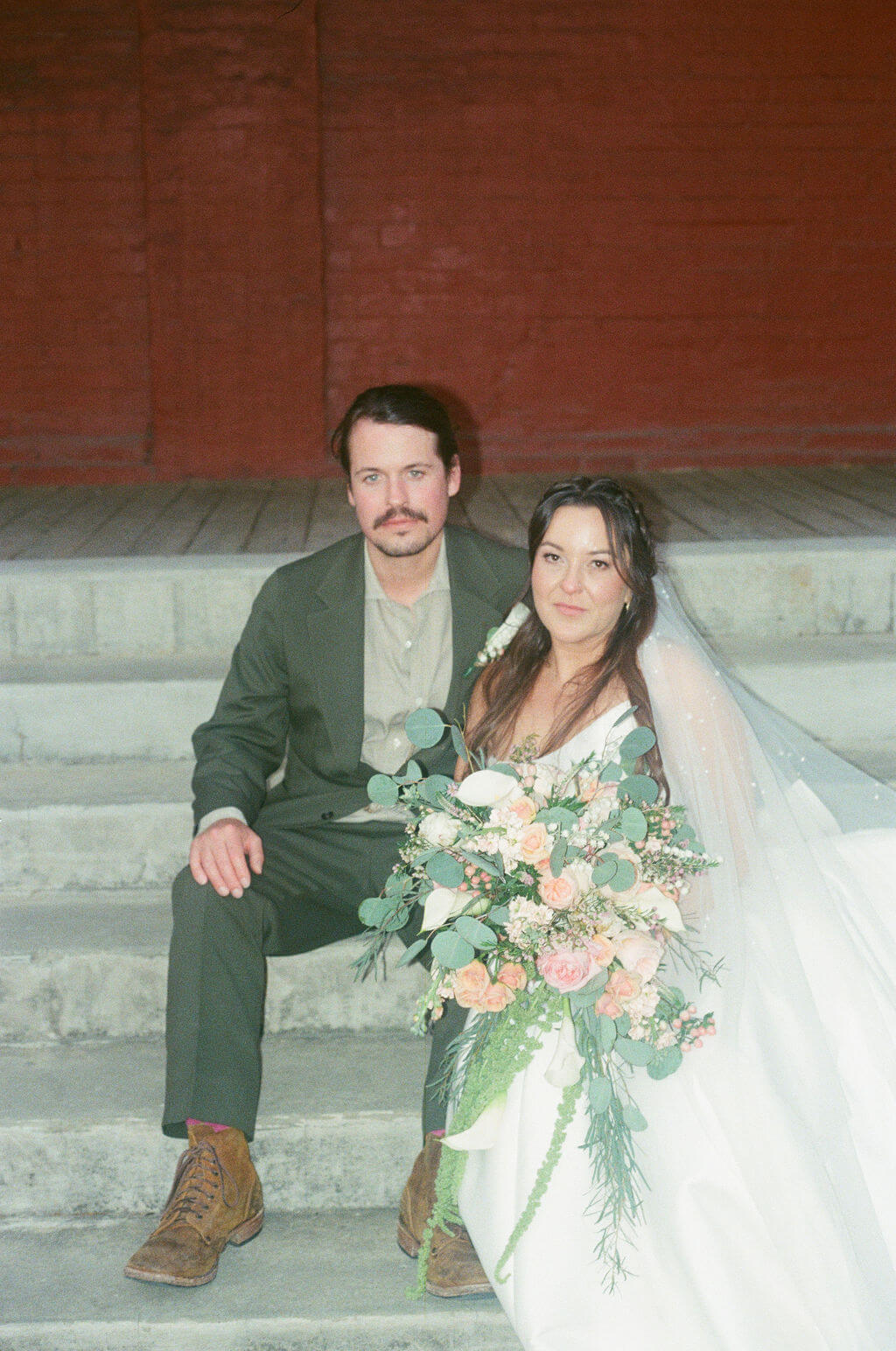 Bride and groom sitting side by side on concrete steps against a red brick wall, the groom in an olive suit and brown boots and the bride holding her bouquet