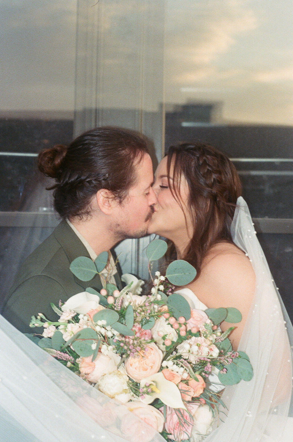 Bride and groom kissing in front of a window with her bouquet of calla lilies, peach roses, and eucalyptus in front