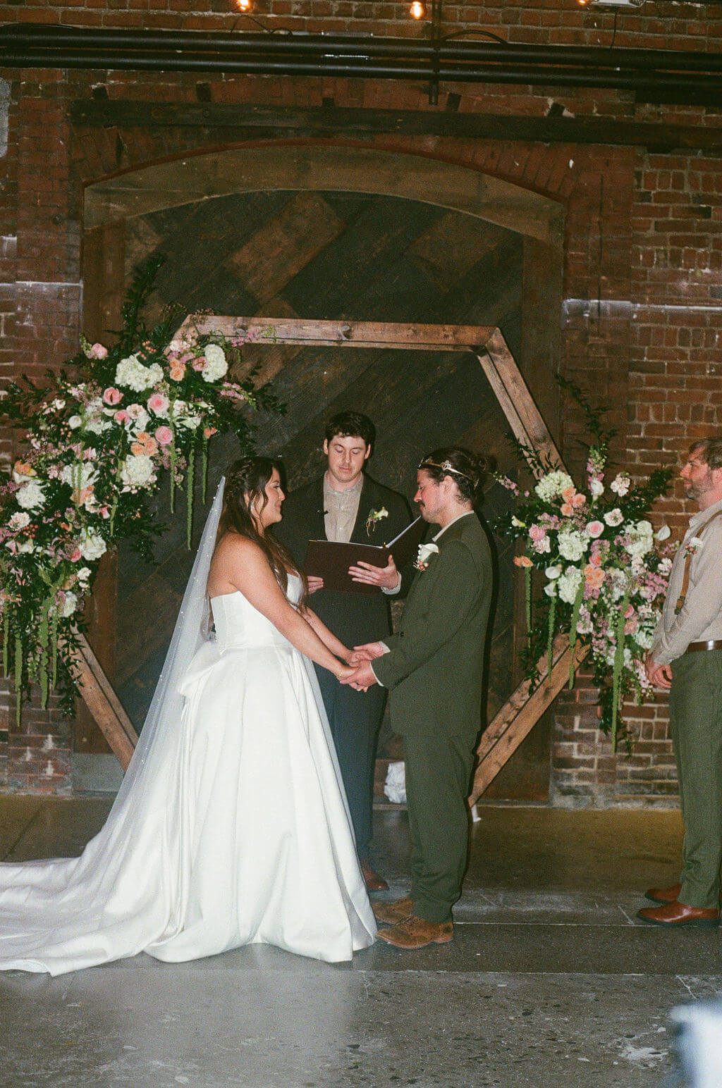 Bride and groom holding hands at the altar in front of a floral hexagon arch with the officiant and groomsman looking on
