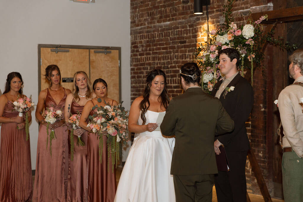 Bride smiling as the groom reads his vows, with bridesmaids in mauve satin dresses holding bouquets beside a floral arrangement and brick wall