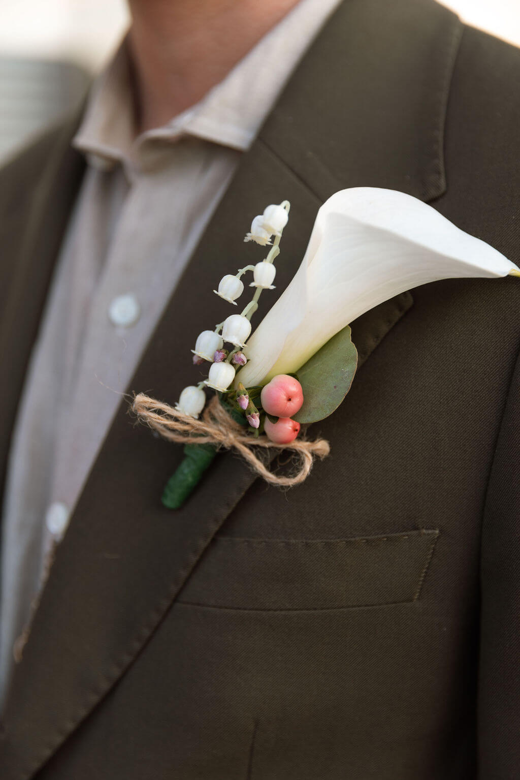 Close-up of the groom's boutonniere with a white calla lily, lily of the valley, pink berries, and twine wrapped stems pinned to an olive suit jacket