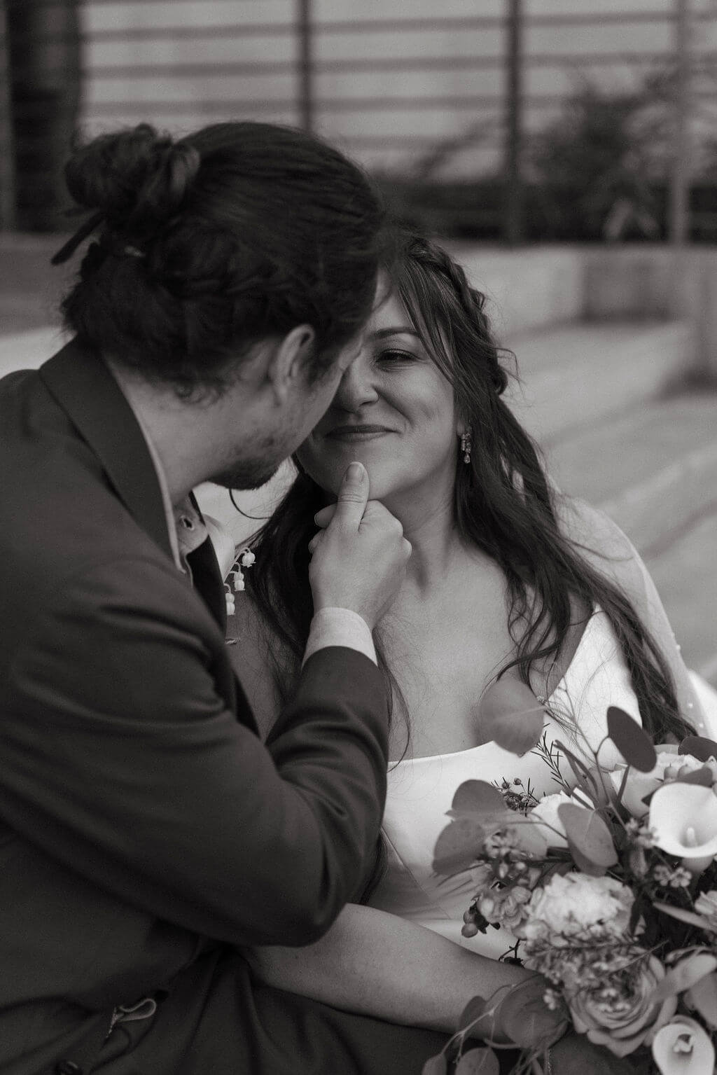 Black and white photo of the bride and groom sitting close together on steps, foreheads nearly touching, with her bouquet in her lap