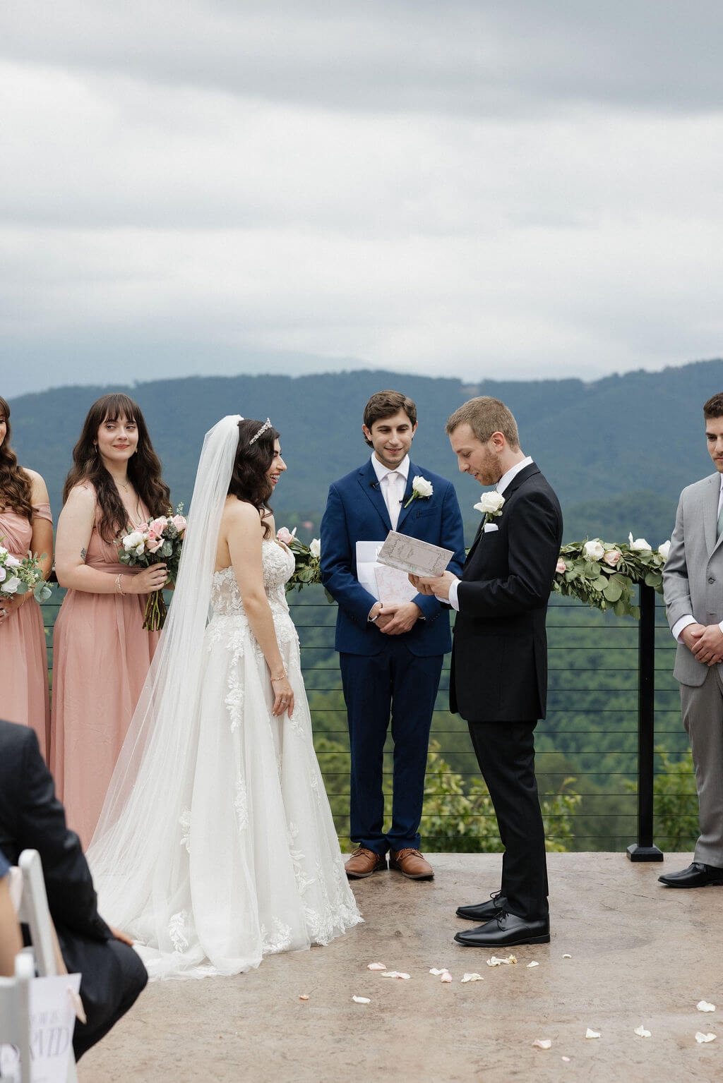 Bride smiling at the groom as he reads his vows during their outdoor wedding ceremony.