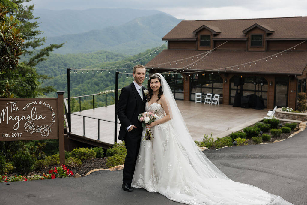 Bride and groom posing in front of The Magnolia wedding venue with sweeping Smoky Mountain views behind them.