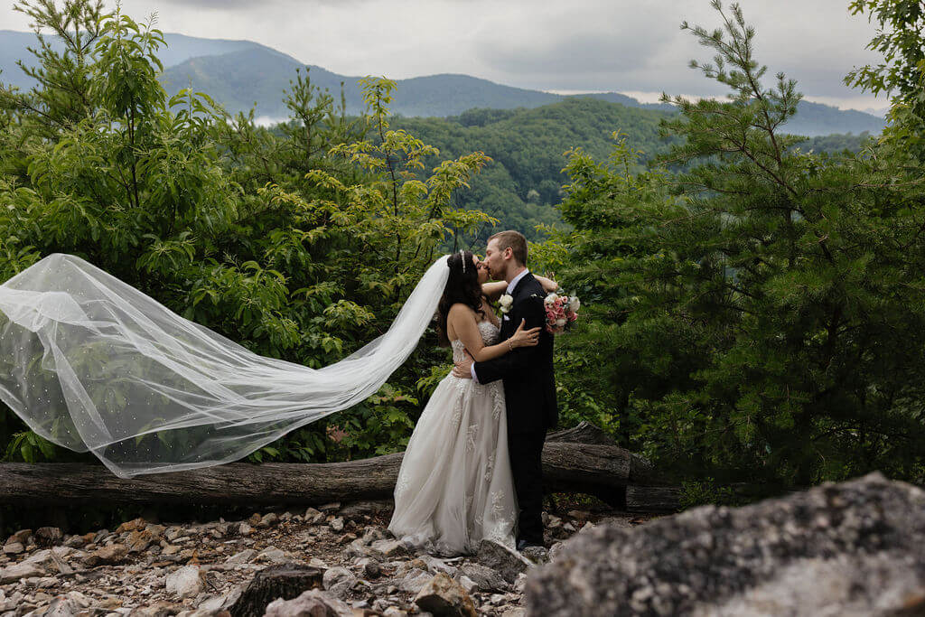 Bride and groom kissing on a rocky Smoky Mountain overlook as the bride’s long veil blows dramatically in the wind.