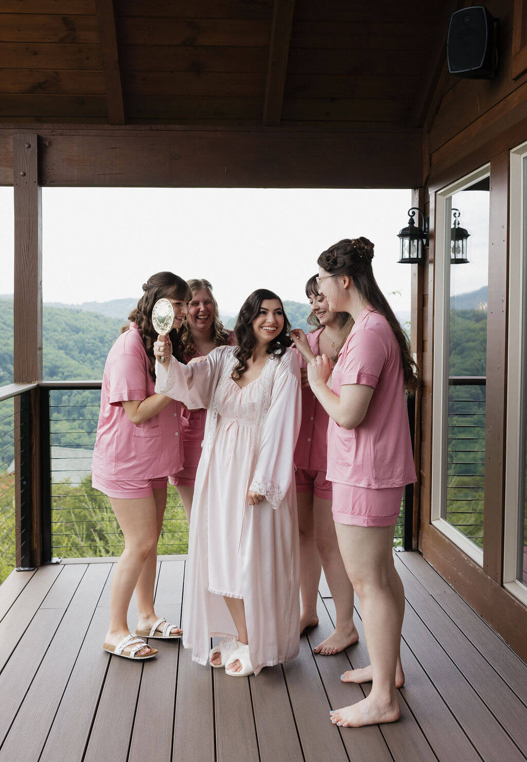 Bride in a white gown and bridesmaids in pink dresses share a moment on a covered porch.