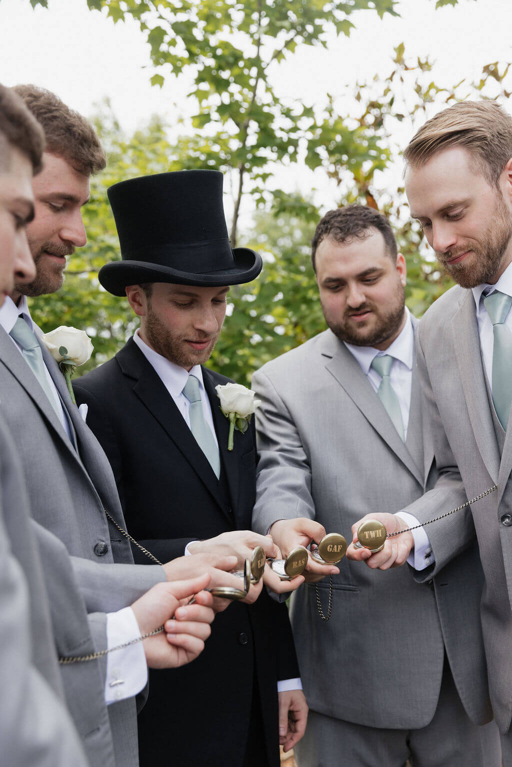 Groom and groomsmen showing matching pocket watches during wedding party portraits.