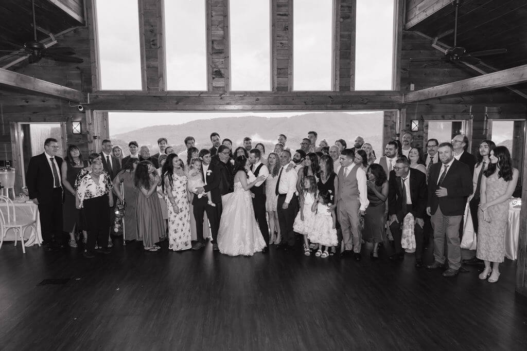 Black-and-white group portrait of wedding guests gathered inside a rustic reception hall.