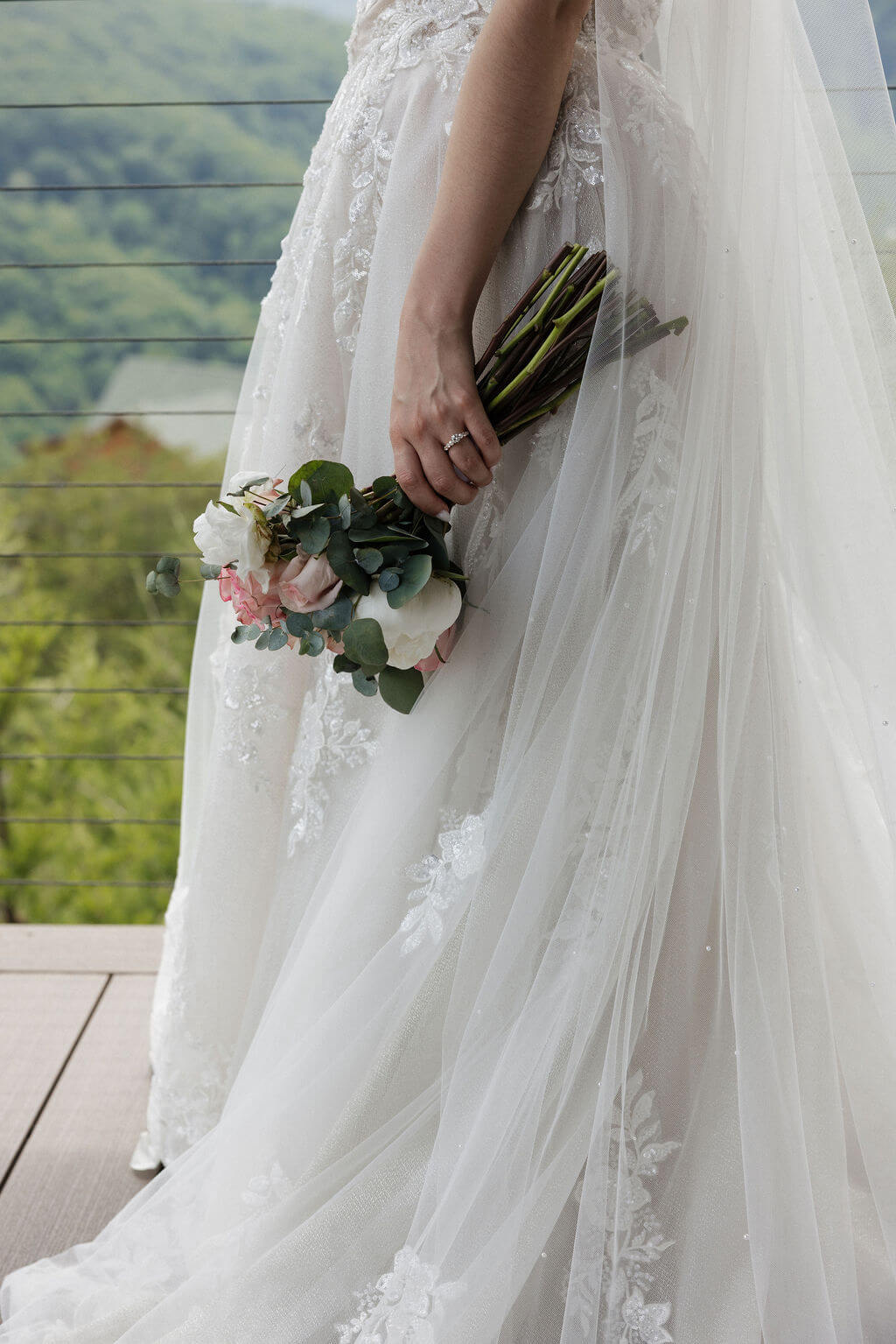 Close-up of the bride’s lace wedding dress, bouquet, and wedding ring with mountain views behind her.