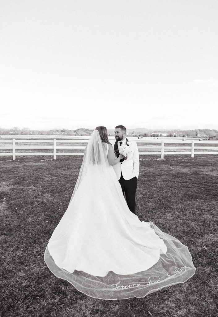 Black-and-white wedding portrait of a bride and groom embracing in an open field, showcasing the bride's long embroidered veil and flowing wedding gown.