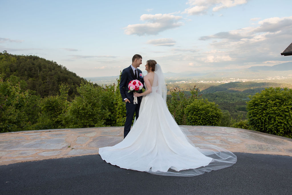 Bride and groom stand together on a stone overlook with a sweeping mountain landscape behind them.