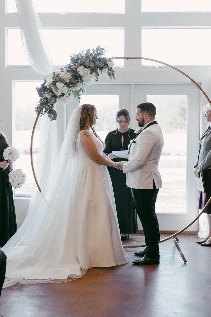 Bride and groom exchanging vows beneath a circular gold wedding arch decorated with white flowers and greenery during an indoor wedding ceremony.