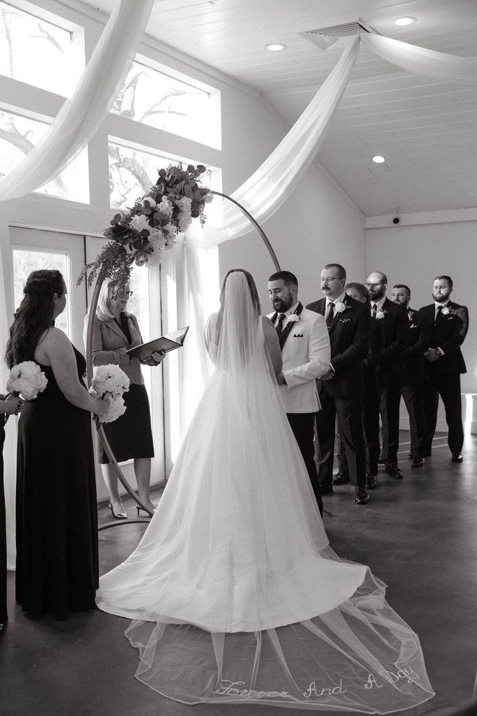 Black-and-white photo of a bride in a flowing wedding gown and long veil standing across from the groom during their indoor wedding ceremony, surrounded by bridesmaids and groomsmen.