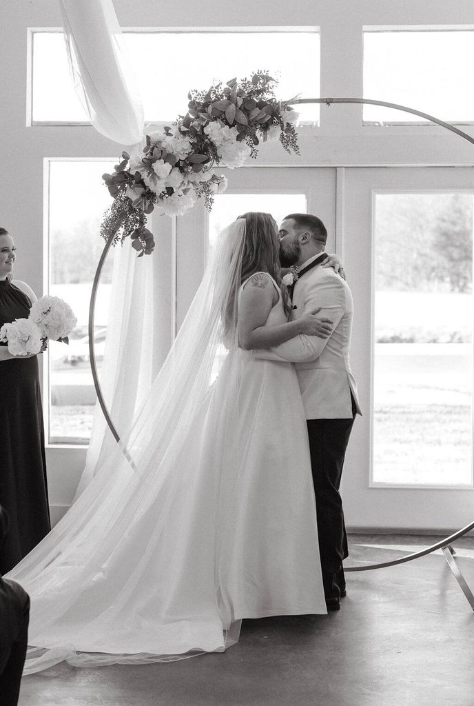 Black-and-white photo of newlyweds kissing beneath a circular floral wedding arch during their indoor ceremony, with the bride's long veil trailing behind her.