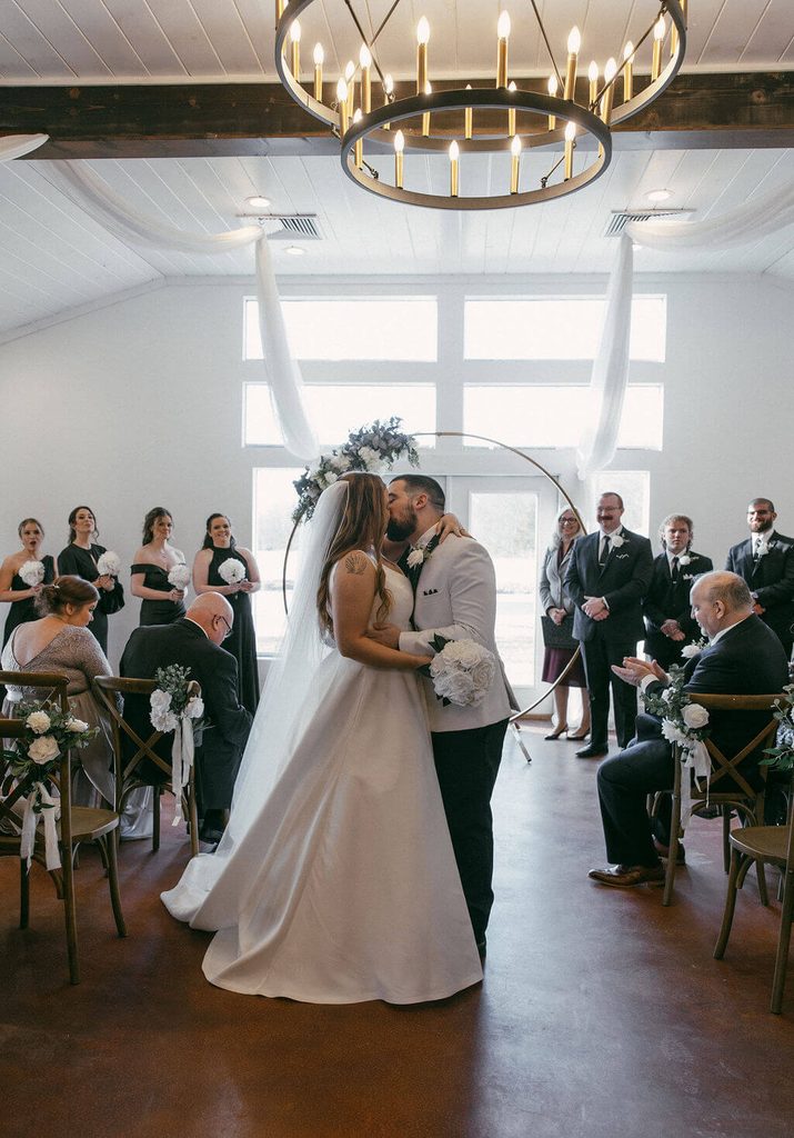 Newlyweds sharing their first kiss beneath a floral wedding arch inside a bright white wedding venue as guests watch from their seats.
